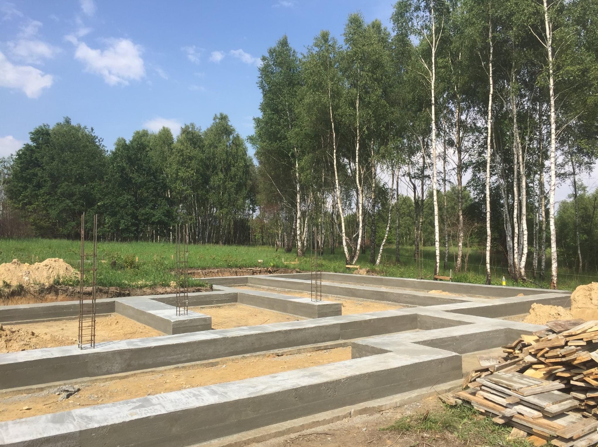 Wylany fundament pod dom jednorodzinny, zbrojenie wystaje pionowo, w tle las brzozowy i błękitne niebo z chmurami, obok stos desek.