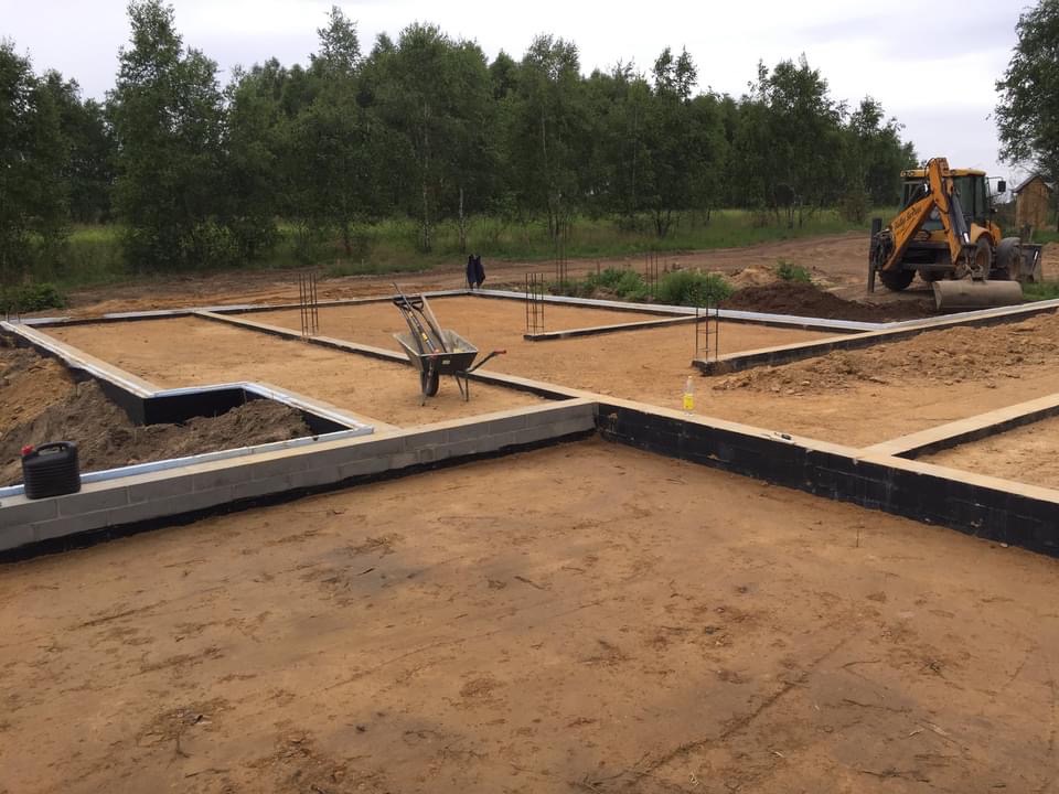 Wykop pod fundamenty domu jednorodzinnego, częściowo wylany beton, widoczna izolacja przeciwwilgociowa, zbrojenie, taczka z narzędziami i koparko-ładowarka w tle.