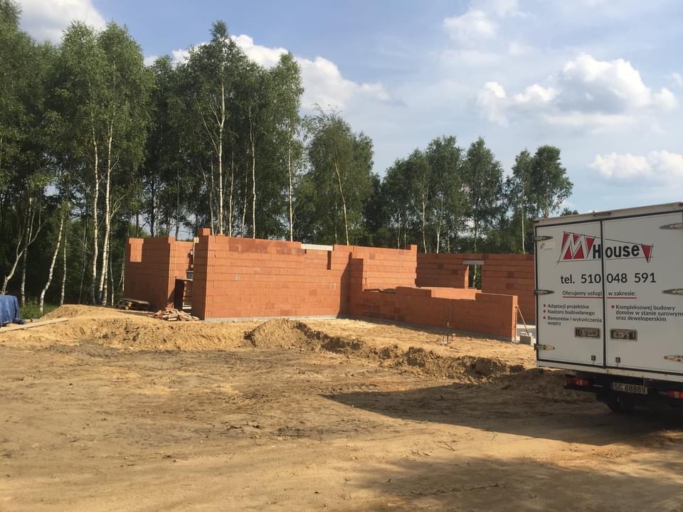 Budowa domu murowanego w stanie surowym otwartym, widoczne ściany z czerwonej cegły, na drugim planie brzozowy lasek i samochód dostawczy z logo firmy budowlanej.