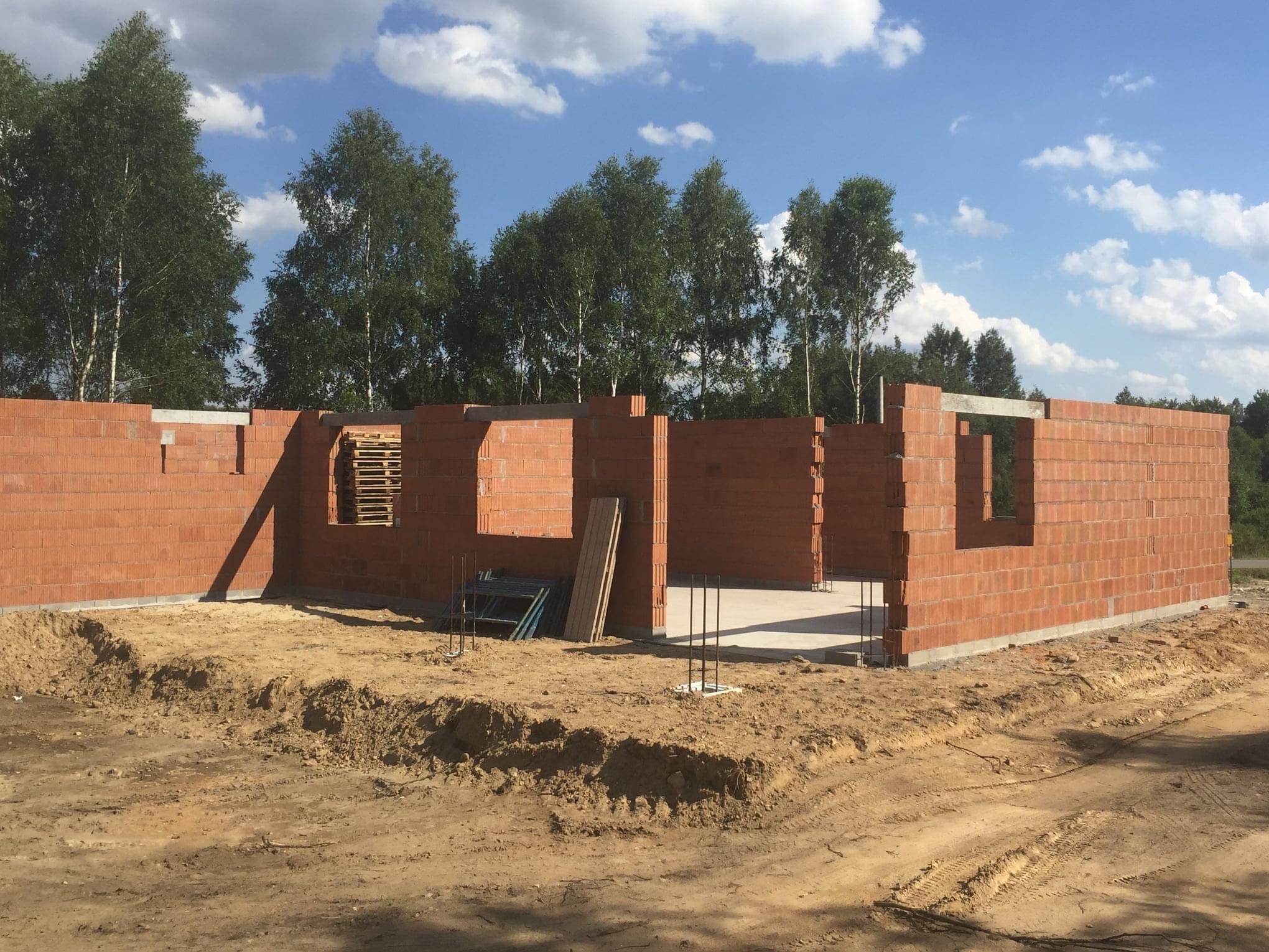 Wznoszenie ścian parteru z czerwonej cegły na fundamencie w trakcie budowy domu jednorodzinnego, widoczne zbrojenia i stemple, w tle drzewa i błękitne niebo z chmurami.