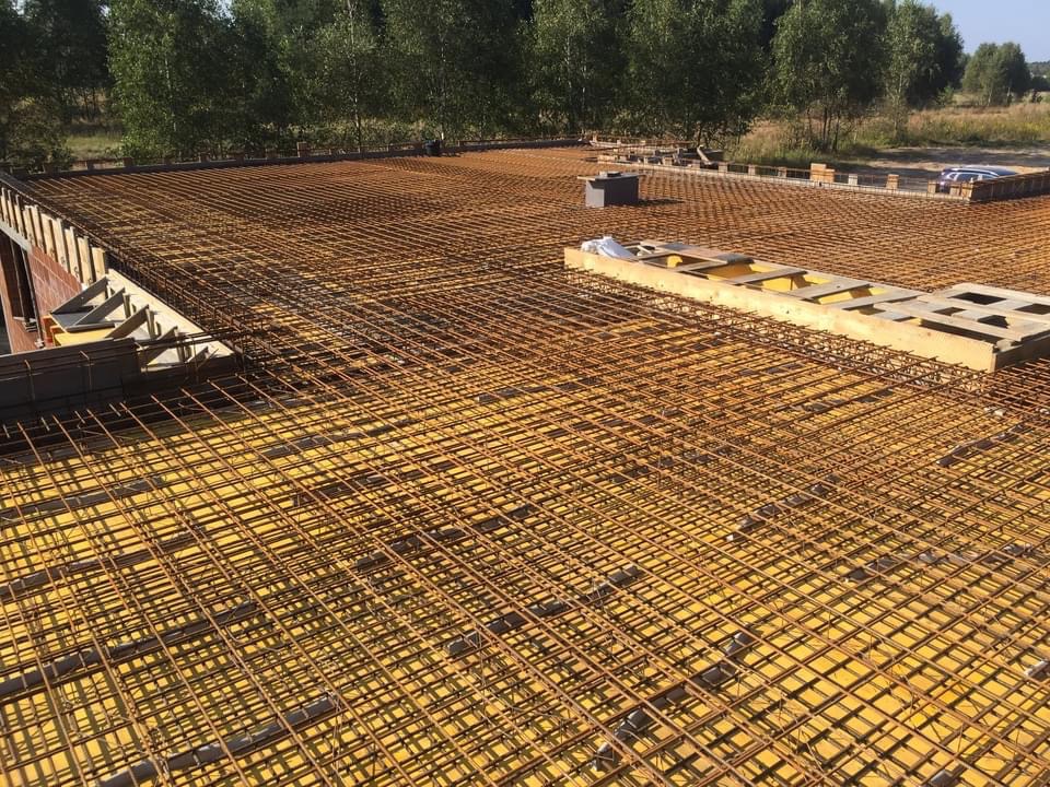 Zbrojenie stropu na żółtej izolacji z widocznymi elementami szalunku i drzewami w tle.