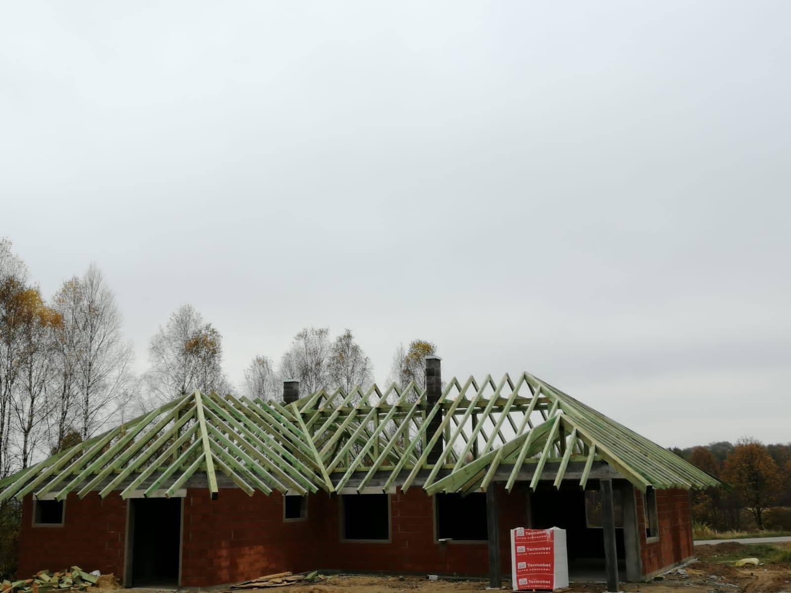 Surowy stan domu z cegły z drewnianą konstrukcją dachu w trakcie budowy, widoczne kominy, na pierwszym planie paczki materiałów budowlanych.