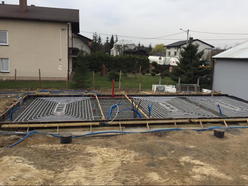 Przygotowanie podłoża pod ogrzewanie podłogowe wewnątrz szalunku fundamentów z widocznymi rurami grzewczymi ułożonymi w pętle, niebieskie rury instalacyjne oraz wystające czerwone rury pionowe,...