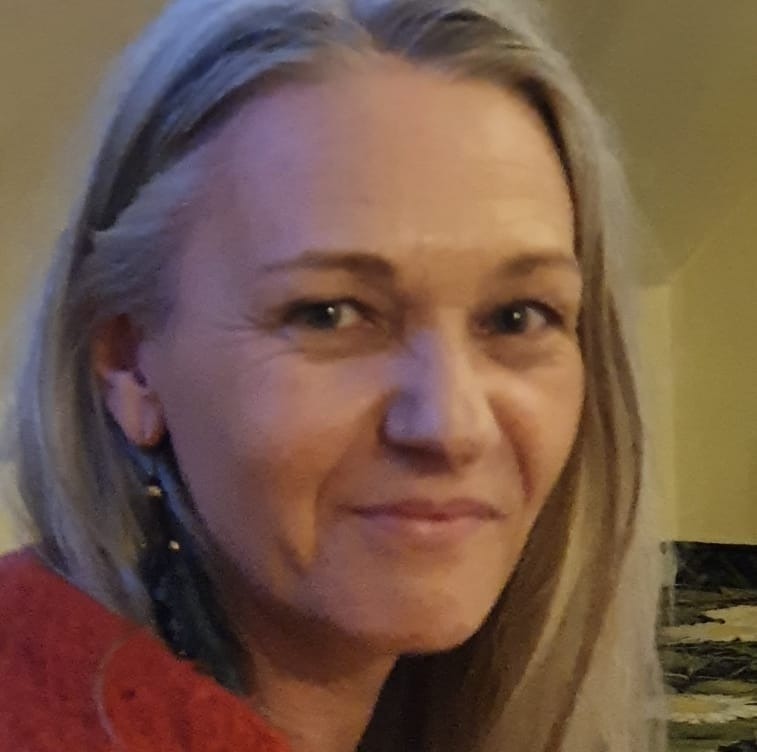 Portret uśmiechniętej kobiety z długimi, siwymi włosami, ubrana w czerwoną bluzkę, z delikatnymi zmarszczkami wokół oczu.