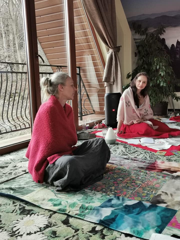 Dwie kobiety siedzą na podłodze w jasnym pomieszczeniu podczas sesji relaksacyjnej, otulone kocami, z dyfuzorem zapachowym w pobliżu, na tle dużych okien i roślin doniczkowych.