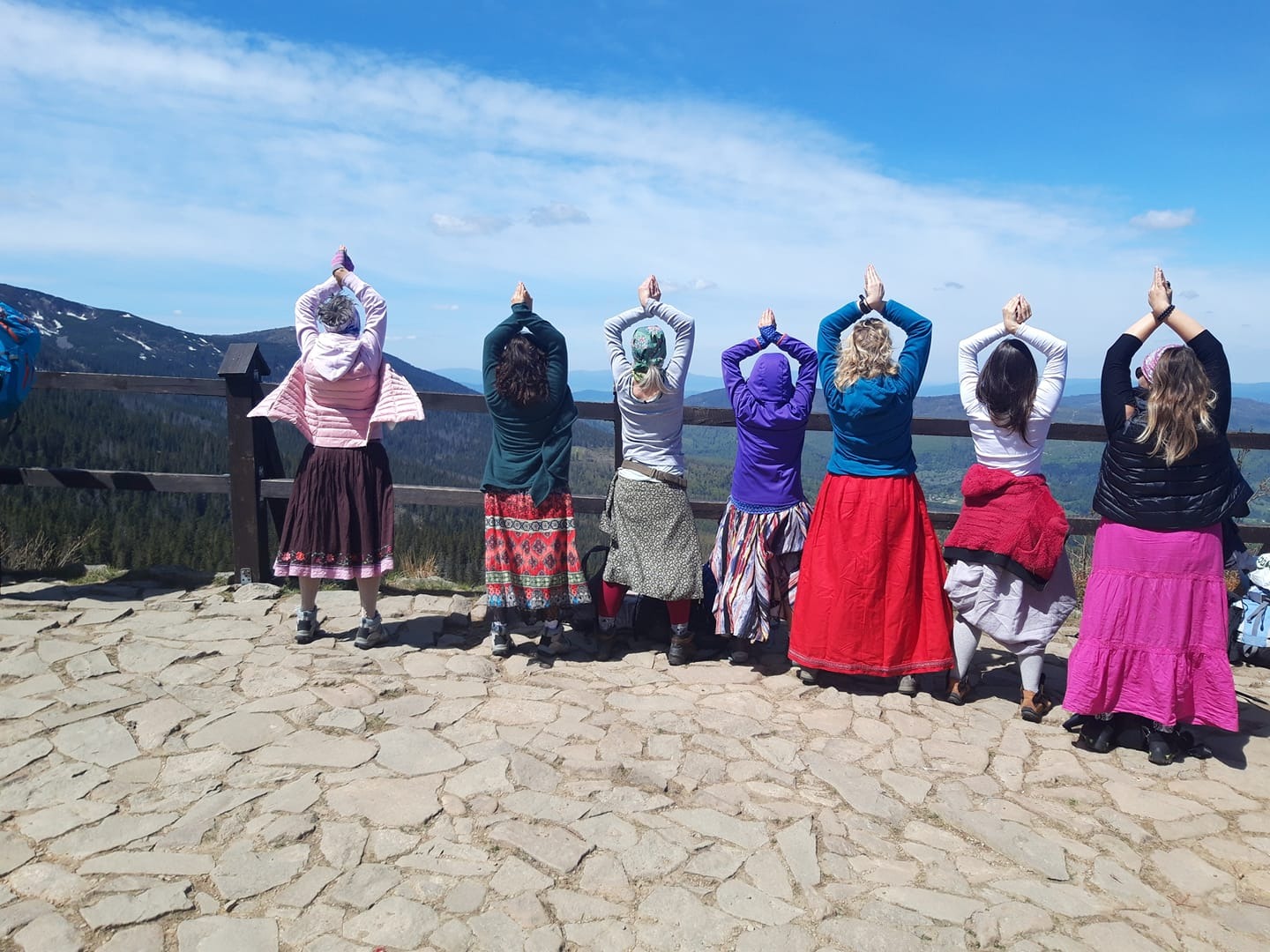 Grupa kobiet tyłem, w długich spódnicach, wykonuje ćwiczenie z rękami uniesionymi do góry na tle górskiego krajobrazu z drewnianą barierką.