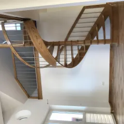 Nowoczesne schody z drewna o jasnym odcieniu z metalowymi elementami balustrady, widok z dołu na zakręcającą konstrukcję w jasnym wnętrzu z oknami.