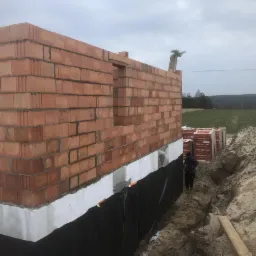 Wznoszenie ścian z czerwonej cegły ceramicznej na fundamencie z izolacją przeciwwilgociową, widoczne palety z cegłami i wykop w gruncie.