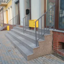 Schody wejściowe do budynku z szarego granitu z metalową balustradą, żółta skrzynka pocztowa zamontowana na ścianie, widok na ulicę.