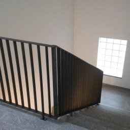 Tadex - Metalowa balustrada schodowa z pionowymi prętami, widok z góry na szare schody i okno z luksferami w tle.