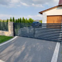 Nowoczesna, pozioma brama wjazdowa w kolorze grafitowym, z automatyką, zamontowana na posesji z kostką brukową i elementami ogrodzenia z kamienia łupanego, na tle domu z drewnianą bramą garażową.