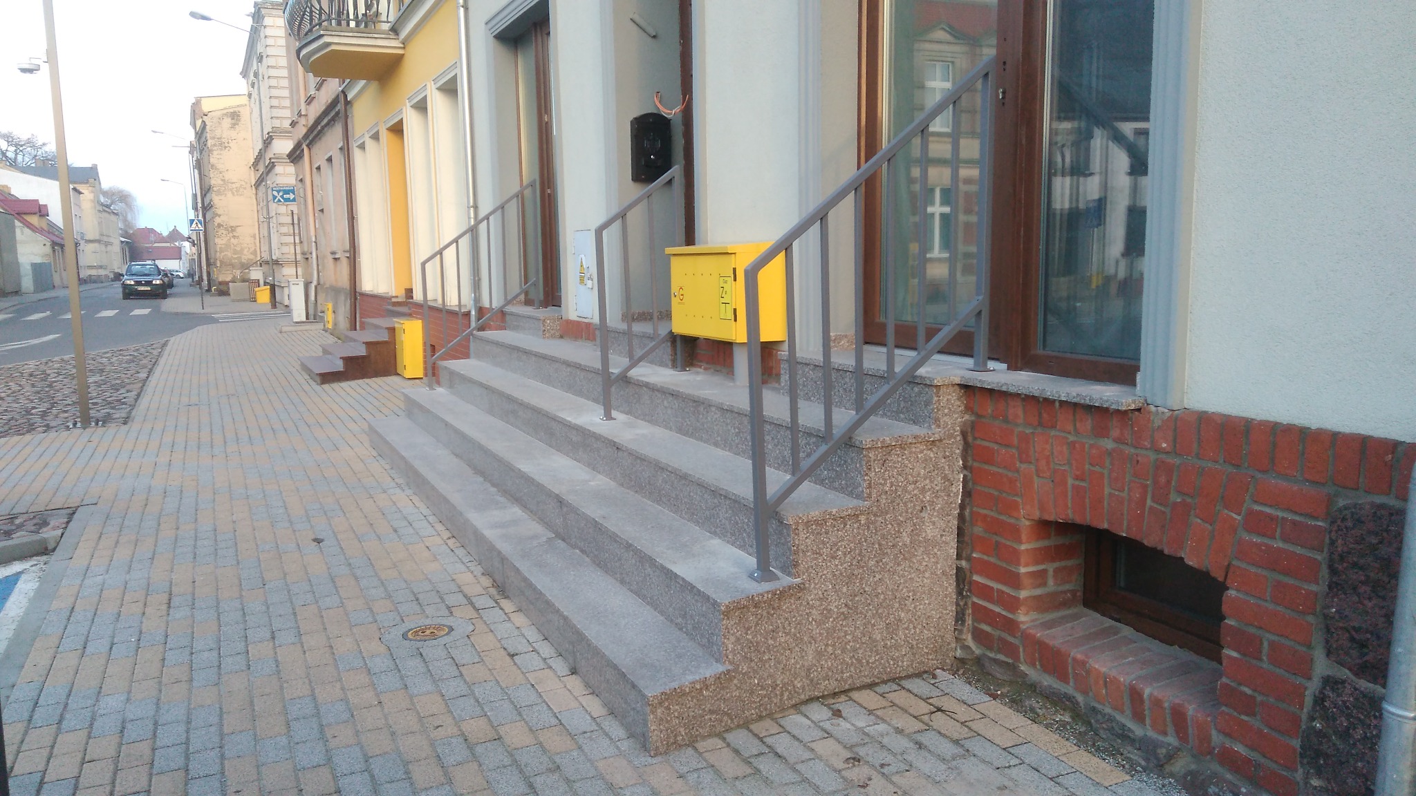 Schody wejściowe do budynku z szarego granitu z metalową balustradą, żółta skrzynka pocztowa zamontowana na ścianie, widok na ulicę.