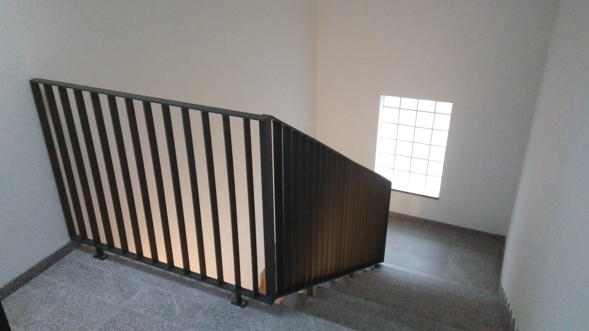 Metalowa balustrada schodowa z pionowymi prętami, widok z góry na szare schody i okno z luksferami w tle.