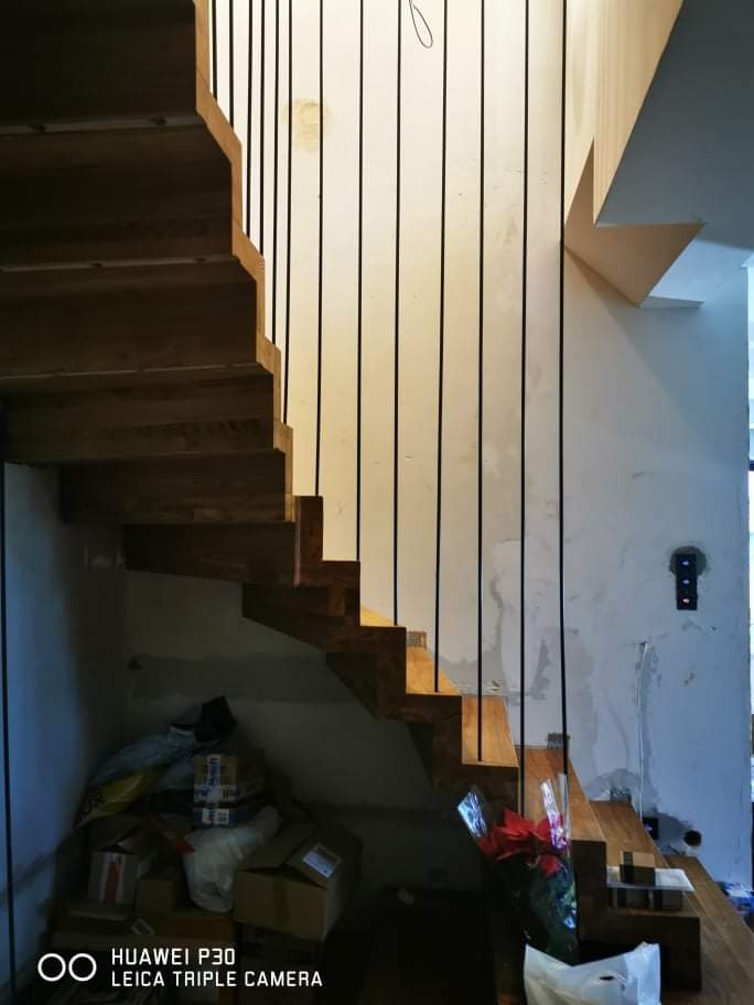 Drewniane schody z czarnymi, pionowymi balustradami, widok z dołu, w tle ściana w trakcie wykończenia i sterta kartonów pod schodami.