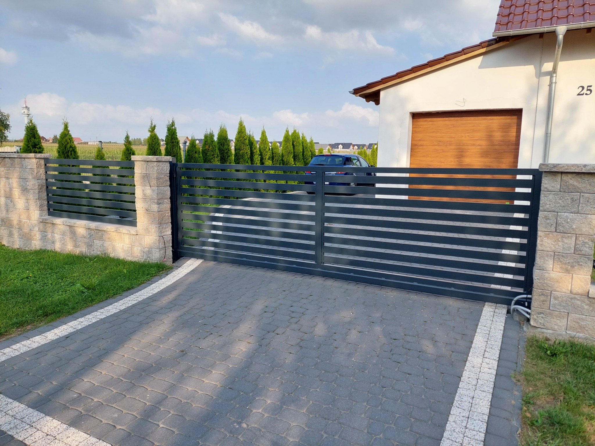 Nowoczesna, pozioma brama wjazdowa w kolorze grafitowym, z automatyką, zamontowana na posesji z kostką brukową i elementami ogrodzenia z kamienia łupanego, na tle domu z drewnianą bramą garażową.
