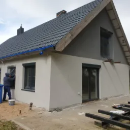 Elewacja domu jednorodzinnego w trakcie prac wykończeniowych, z widocznym pracownikiem malującym okolice okna, zabezpieczonego folią. Część ściany pomalowana na szaro, dolna na beżowo. Na pierwszym...