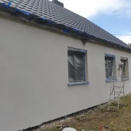 Elewacja domu jednorodzinnego w trakcie malowania natryskowego, okna zabezpieczone folią, drabina oparta o ścianę, widoczny pistolet do malowania.