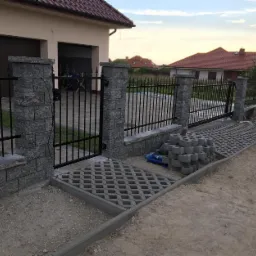 Czarny metalowy płot z bramą i szarymi kamiennymi słupkami w trakcie montażu, widoczne ułożone płyty ażurowe i stos kamieni.