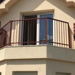 Balkon z metalową balustradą w kolorze bordowym, widok z zewnątrz, jasna elewacja budynku, brązowa dachówka.
