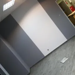 Świeżo pomalowane biuro z ukośnym podziałem kolorów szarości i bieli na ścianie, podłoga wyłożona szarą imitacją drewna, widoczna apteczka pierwszej pomocy na ścianie oraz fragment pomieszczenia...