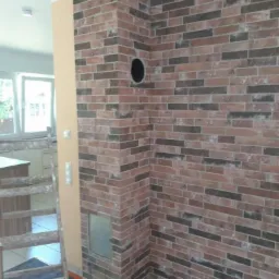 Ściana wykończona cegłą klinkierową z otworem wentylacyjnym, widoczny fragment drabiny i elementy kuchni w tle.