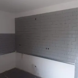 Ściana w trakcie układania szarych, prostokątnych płytek ceramicznych w kuchni, widoczne puszki elektryczne i metalowy profil montażowy.