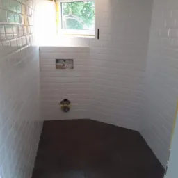 Wnętrze łazienki w trakcie prac wykończeniowych, z białymi, błyszczącymi płytkami ułożonymi w cegiełkę na ścianach, świeżo położona ciemna podłoga, widoczne instalacje wodne i okno z widokiem...