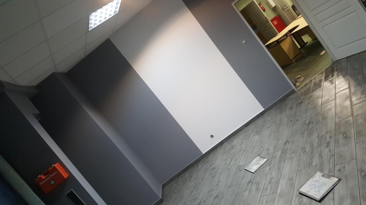 Świeżo pomalowane biuro z ukośnym podziałem kolorów szarości i bieli na ścianie, podłoga wyłożona szarą imitacją drewna, widoczna apteczka pierwszej pomocy na ścianie oraz fragment pomieszczenia...