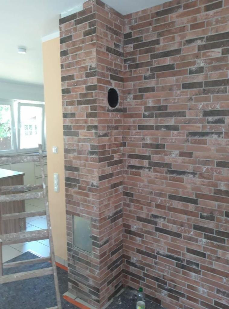 Ściana wykończona cegłą klinkierową z otworem wentylacyjnym, widoczny fragment drabiny i elementy kuchni w tle.