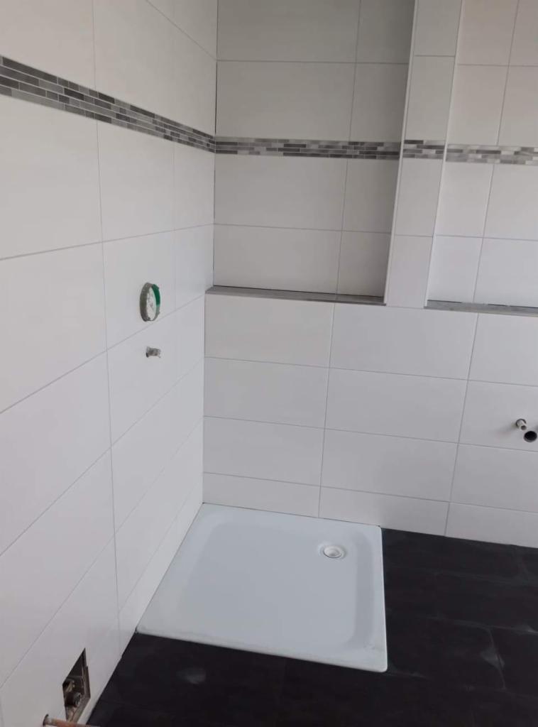 Białe płytki ścienne z szarym pasem dekoracyjnym w wykończonej łazience, widoczny brodzik i ciemna podłoga, przygotowane otwory na armaturę.