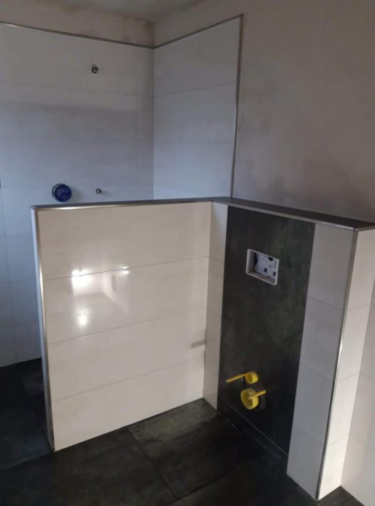 Łazienka w trakcie wykańczania: białe, poziome płytki na ścianach, ciemna, pionowa wnęka z instalacją podtynkową, widoczne elementy instalacji wodno-kanalizacyjnej, ciemna podłoga.