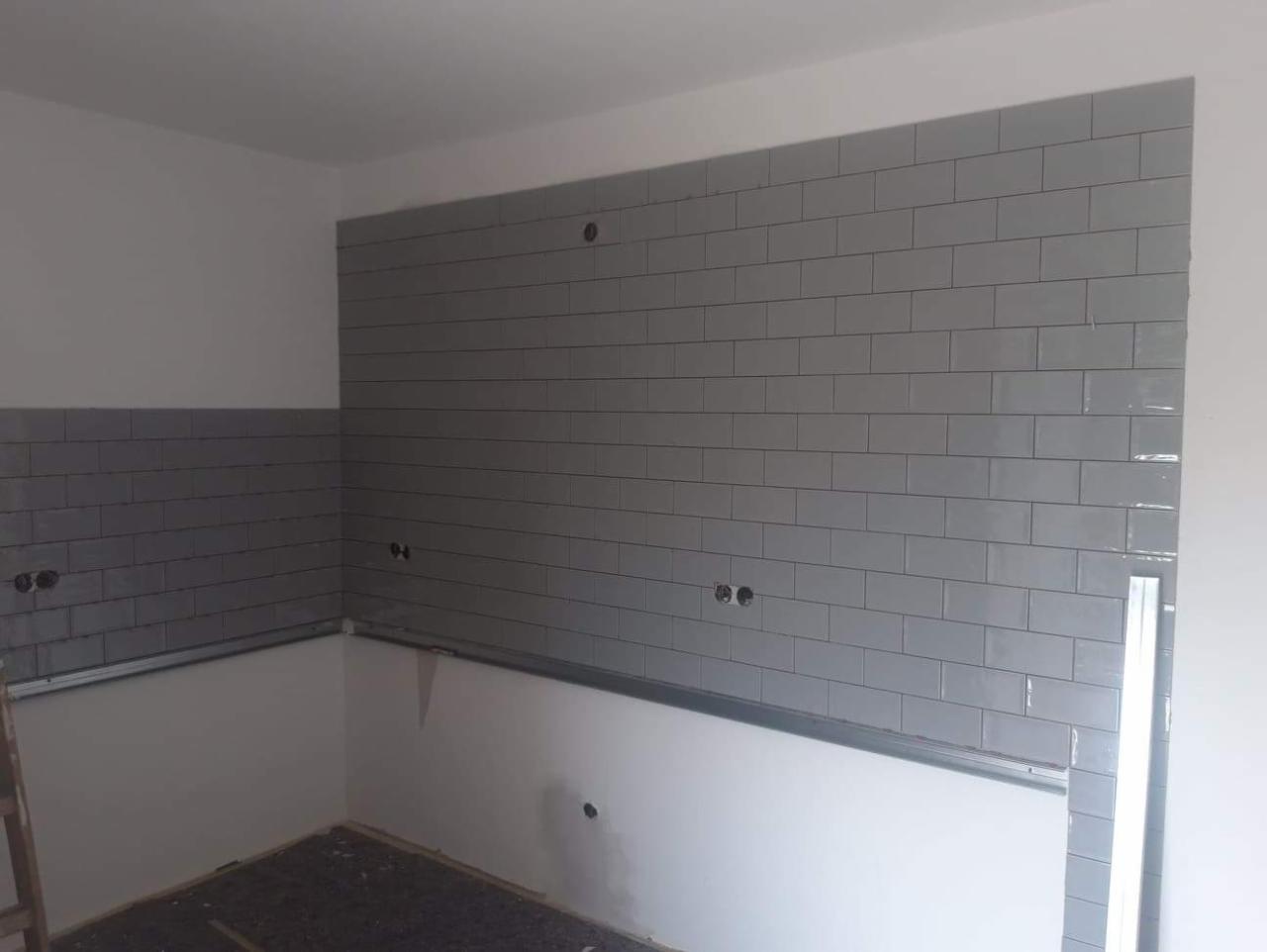 Ściana w trakcie układania szarych, prostokątnych płytek ceramicznych w kuchni, widoczne puszki elektryczne i metalowy profil montażowy.
