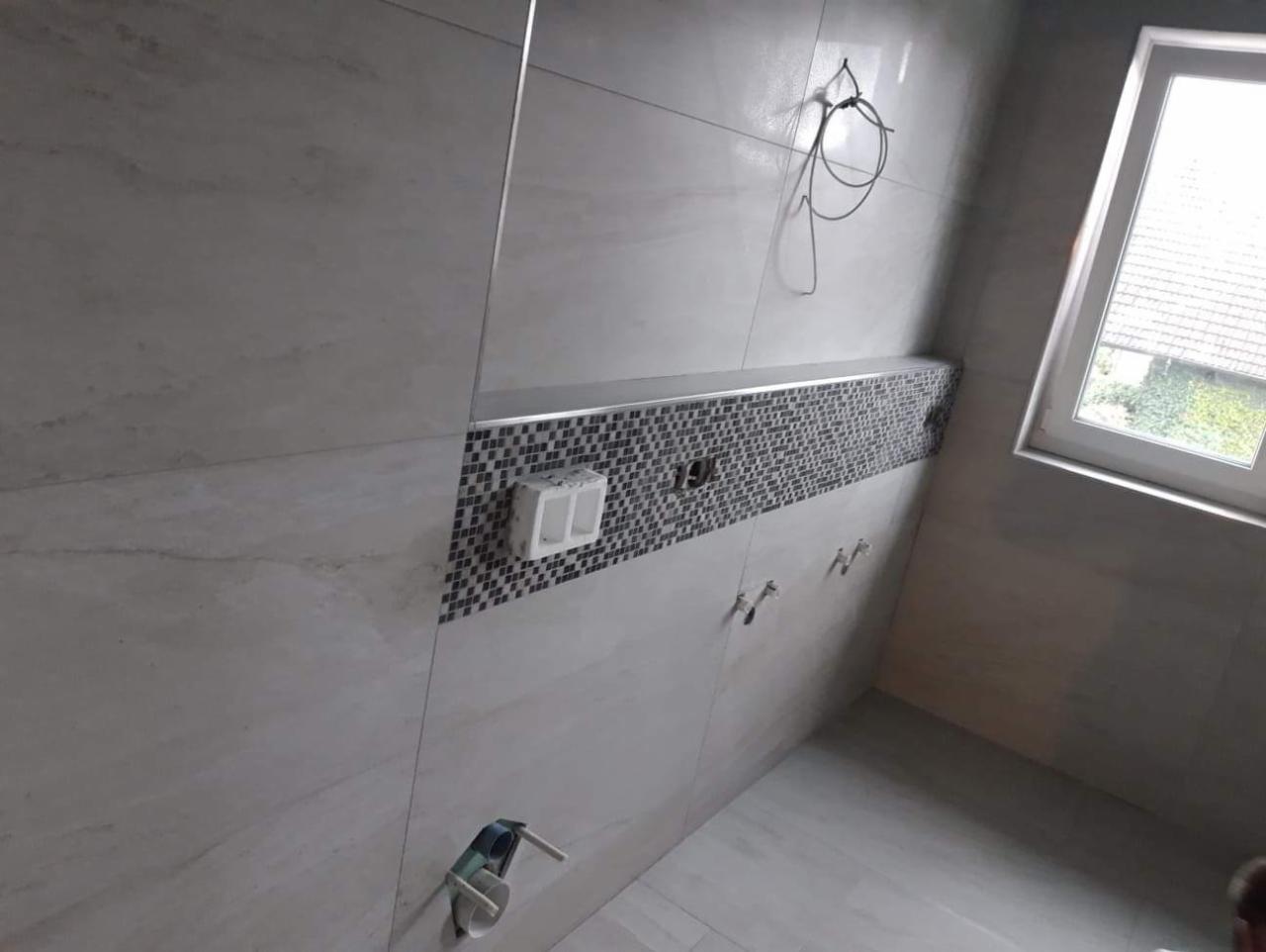 Ściany w łazience wyłożone jasnymi płytkami z dekoracyjnym pasem mozaiki, widoczne przygotowane punkty instalacyjne i okno z widokiem na zieleń.