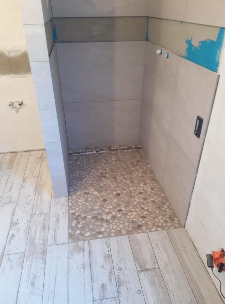 Niedokończona kabina prysznicowa z jasnymi płytkami ściennymi i podłogą wyłożoną drobnymi, naturalnymi kamieniami, widoczna instalacja wodna i częściowo zabezpieczone ściany przed położeniem płytek.