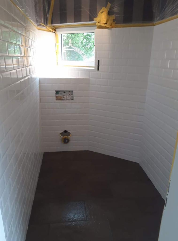 Wnętrze łazienki w trakcie prac wykończeniowych, z białymi, błyszczącymi płytkami ułożonymi w cegiełkę na ścianach, świeżo położona ciemna podłoga, widoczne instalacje wodne i okno z widokiem...