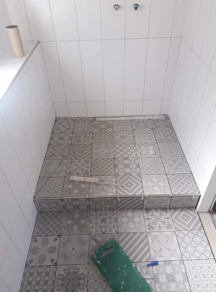 Układanie wzorzystych, szarych płytek podłogowych w małej łazience, widoczne poziomica i narzędzia glazurnicze, białe płytki na ścianach.