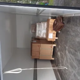 Wnętrze busa z ładunkiem kartonowych pudeł na paletach oraz brązowa walizka na kółkach, widoczne mocowania do zabezpieczenia ładunku.