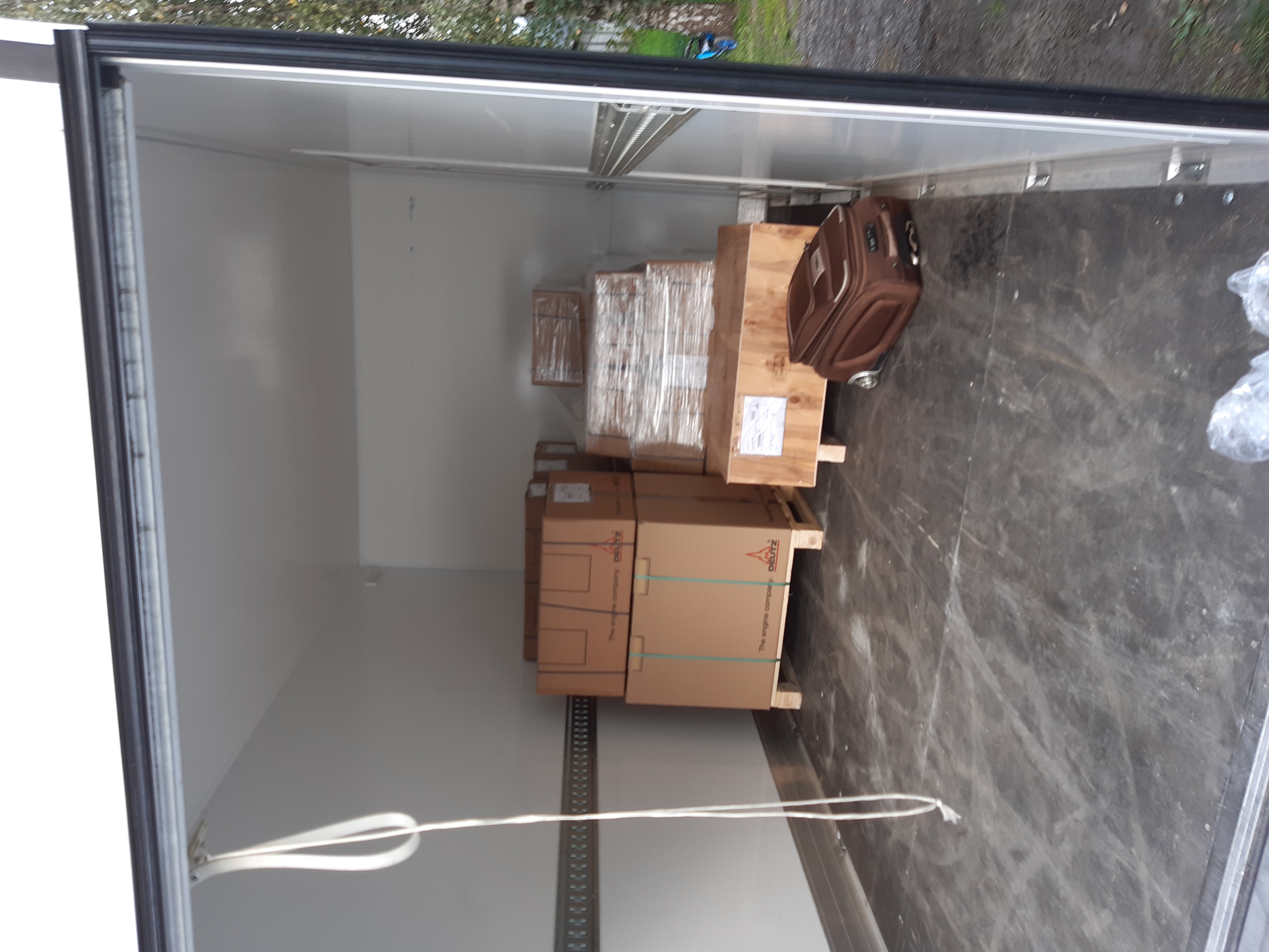 Wnętrze busa z ładunkiem kartonowych pudeł na paletach oraz brązowa walizka na kółkach, widoczne mocowania do zabezpieczenia ładunku.