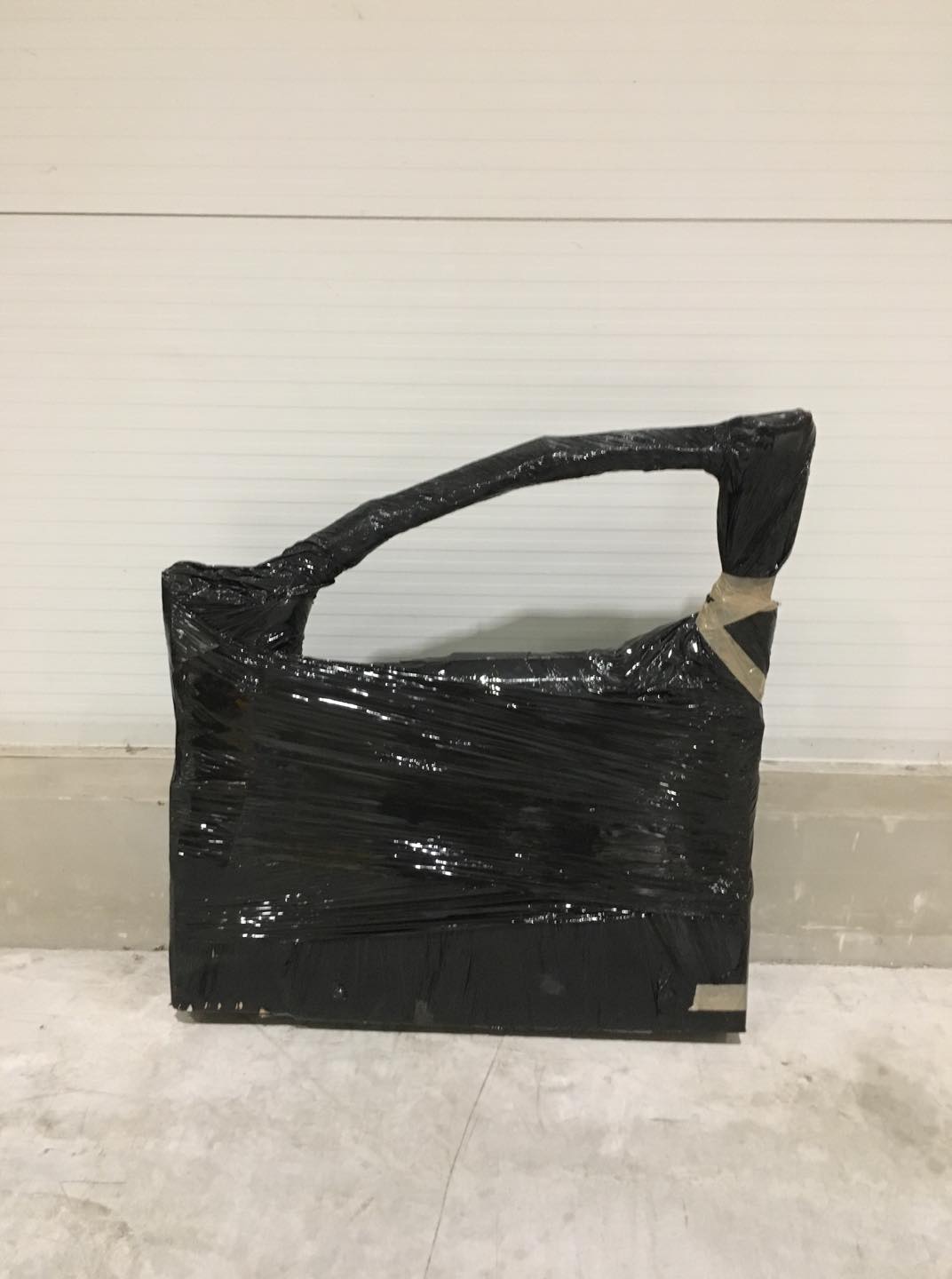 Drzwi samochodowe owinięte czarną folią stretch i zabezpieczone taśmą, gotowe do transportu, na tle białej ściany i betonowej podłogi.