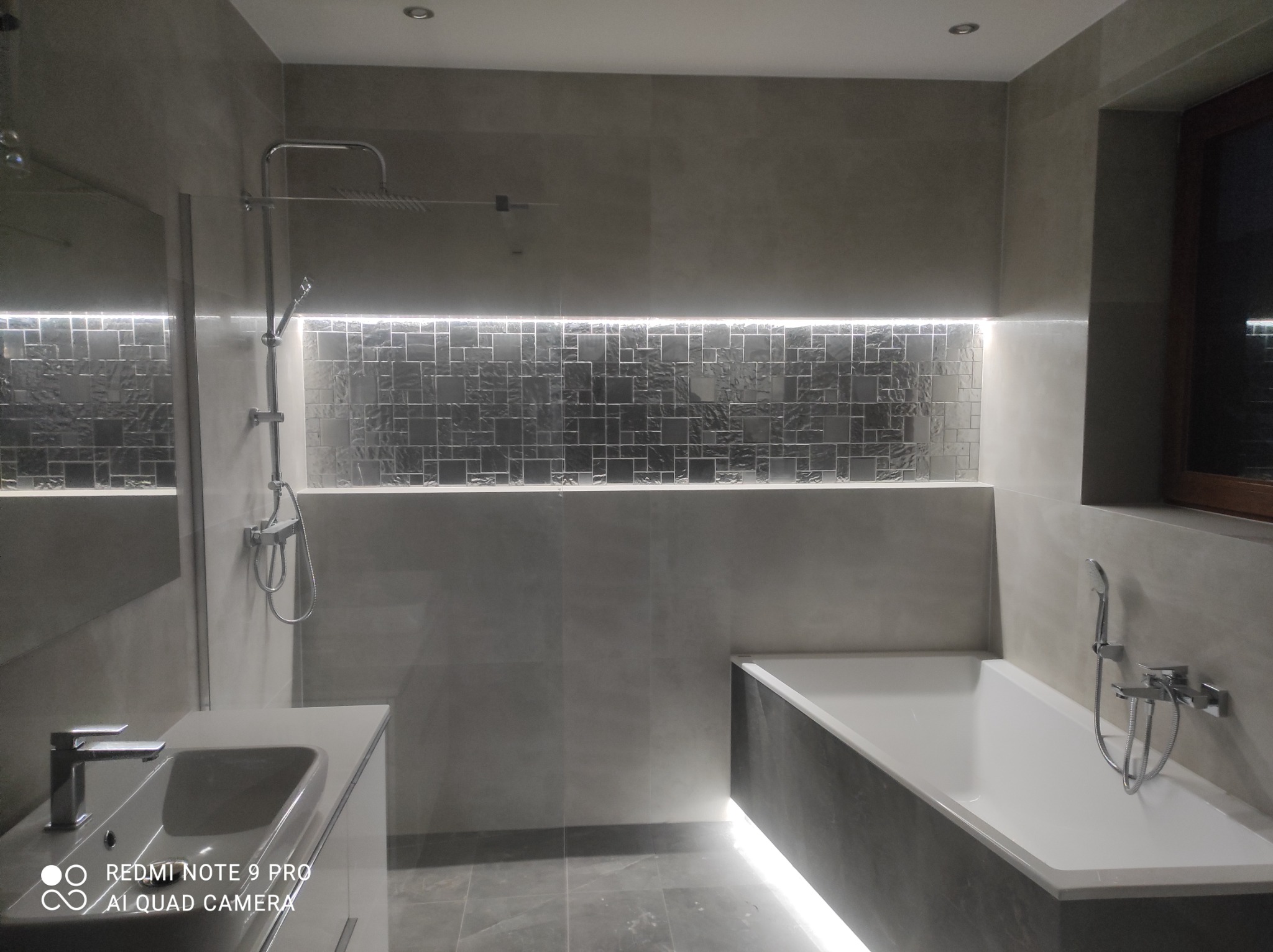 Nowoczesna łazienka z szarymi płytkami, podświetlanymi mozaikami i wanną z podświetleniem LED, minimalistyczny design.