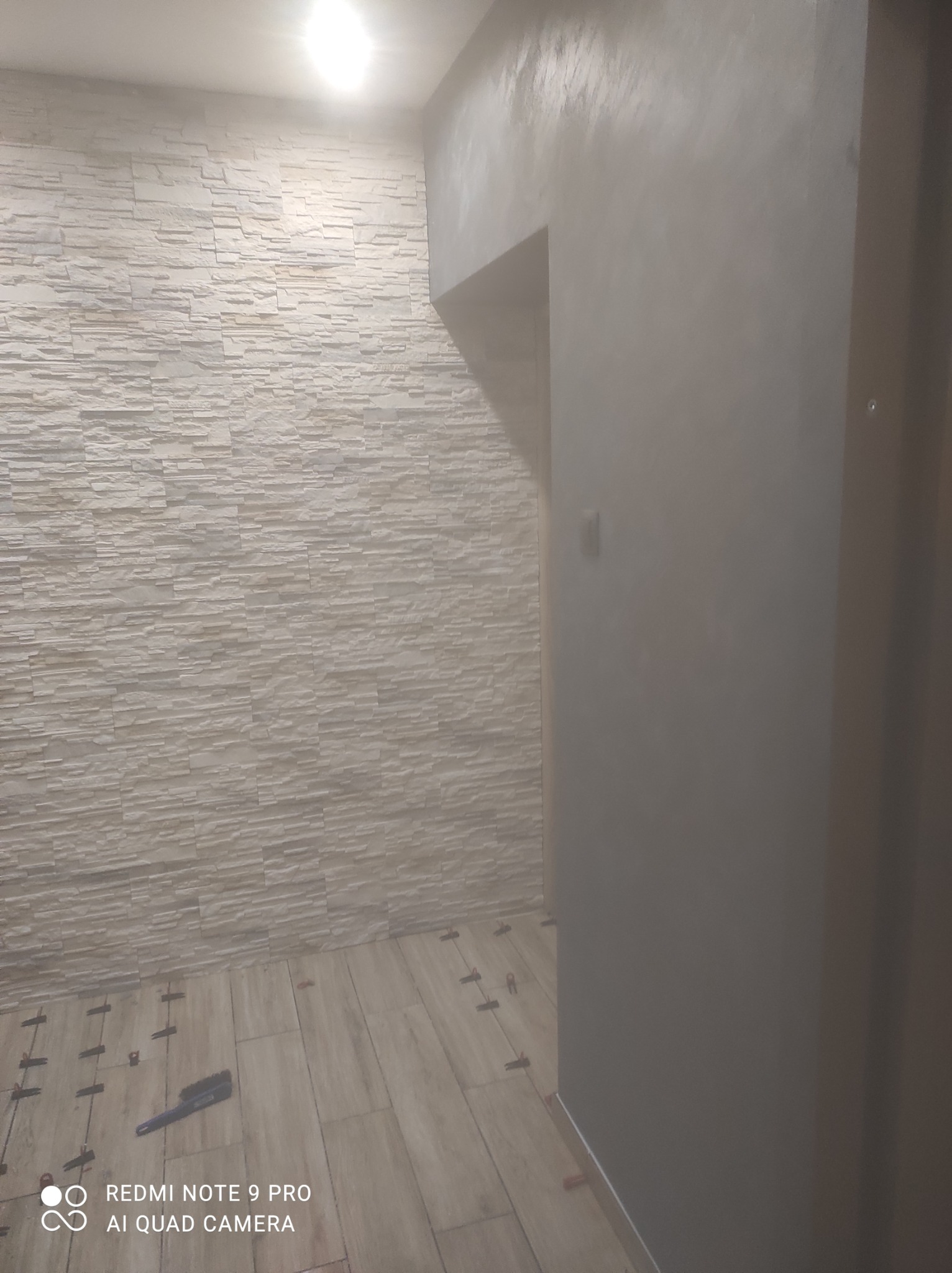 Wnętrze w trakcie remontu: ściana wykończona dekoracyjną okładziną imitującą kamień, sąsiadująca z gładką ścianą, na podłodze płytki z plastikowymi krzyżykami dystansowymi i narzędzie do fugowania.