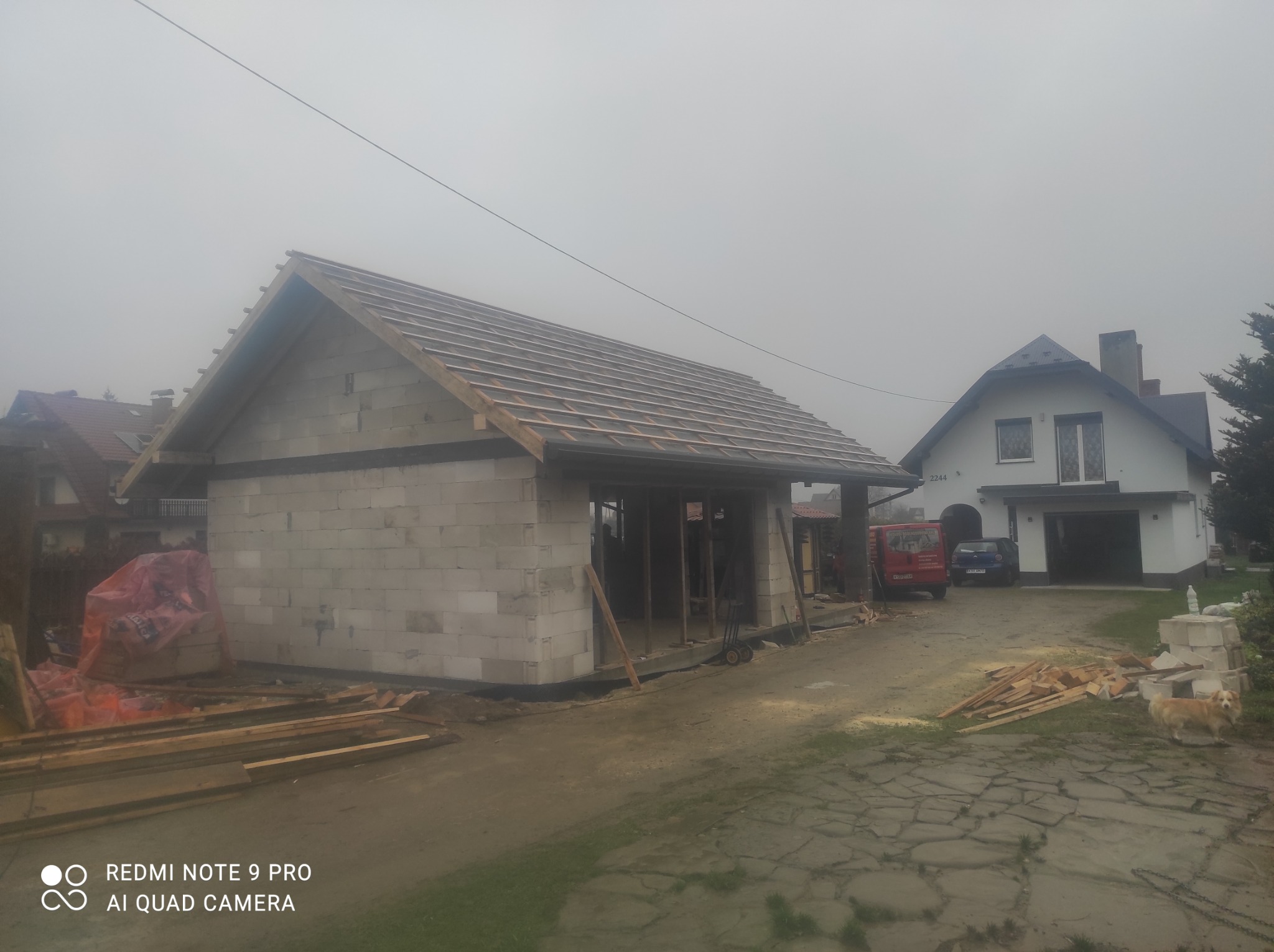 Budowa parterowego budynku z pustaków, widoczna konstrukcja dachu z łatami, w tle dom jednorodzinny i samochody na posesji, pochmurny dzień.