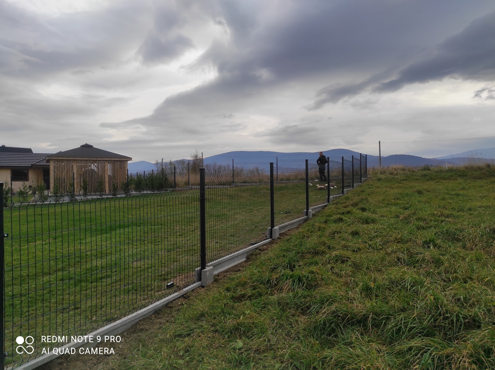 Montaż ogrodzenia panelowego na nierównym terenie, widoczny pracownik w oddali, betonowa podmurówka, trawa, w tle góry i pochmurne niebo.