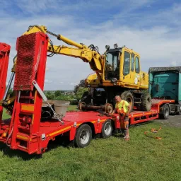 Żółta koparka kołowa przewożona na czerwonej lawecie przez ciężarówkę w Elblągu, operator w odblaskowej kamizelce kontroluje zabezpieczenie ładunku.