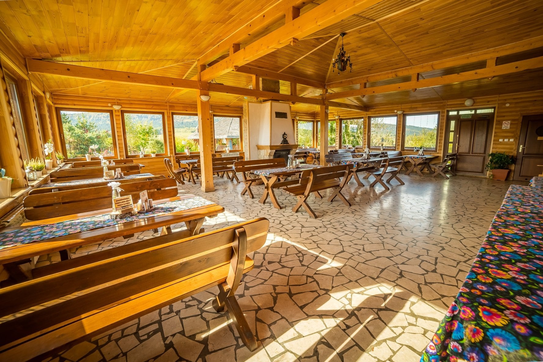 Wnętrze karczmy z drewnianymi ławami i stołami nakrytymi kolorowymi obrusami, kamienna podłoga, duże okna z widokiem na zewnątrz, oświetlenie słoneczne i wiszący żyrandol.