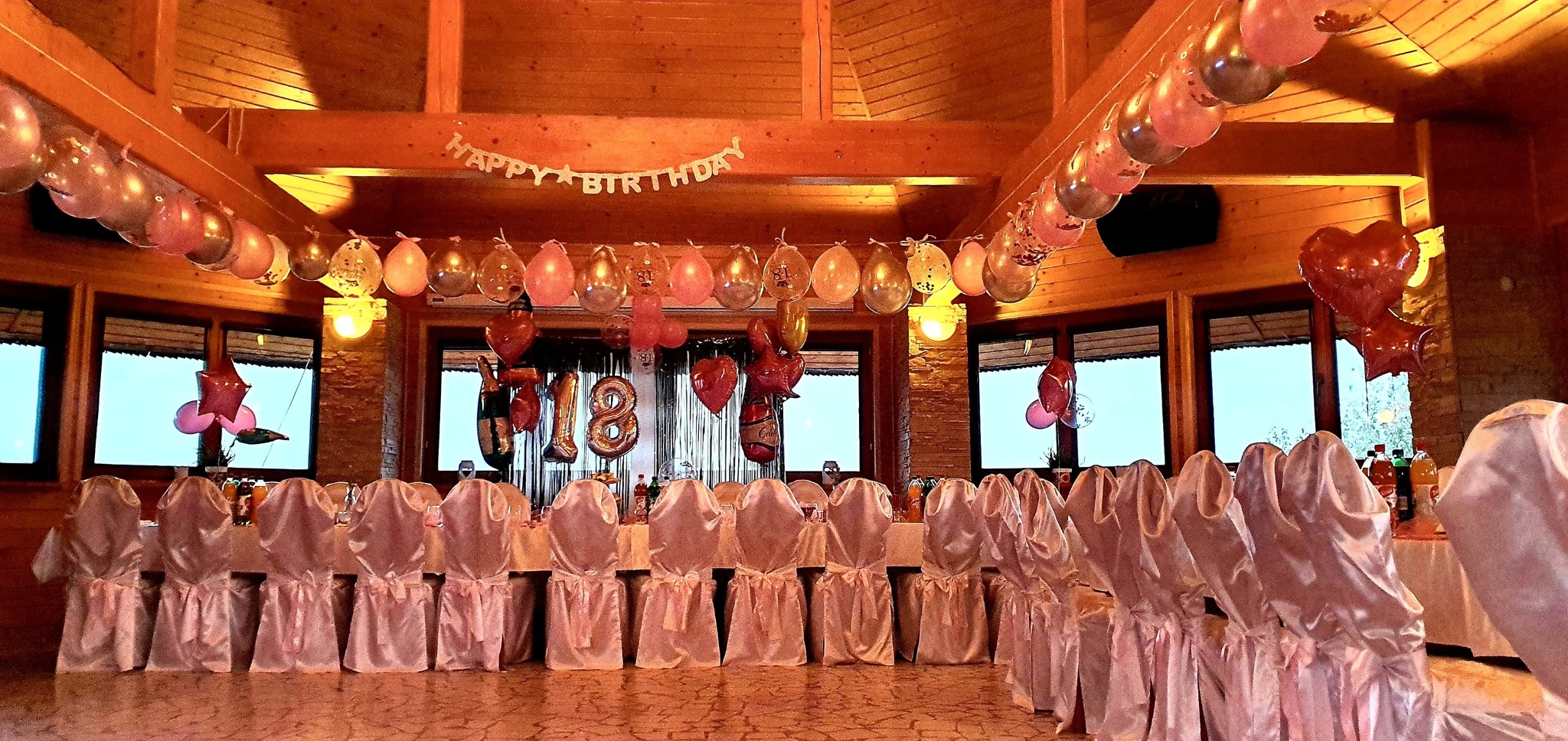 Sala udekorowana na 18 urodziny, z balonami, napisem 'Happy Birthday' i krzesłami w białych pokrowcach z różowymi kokardami.