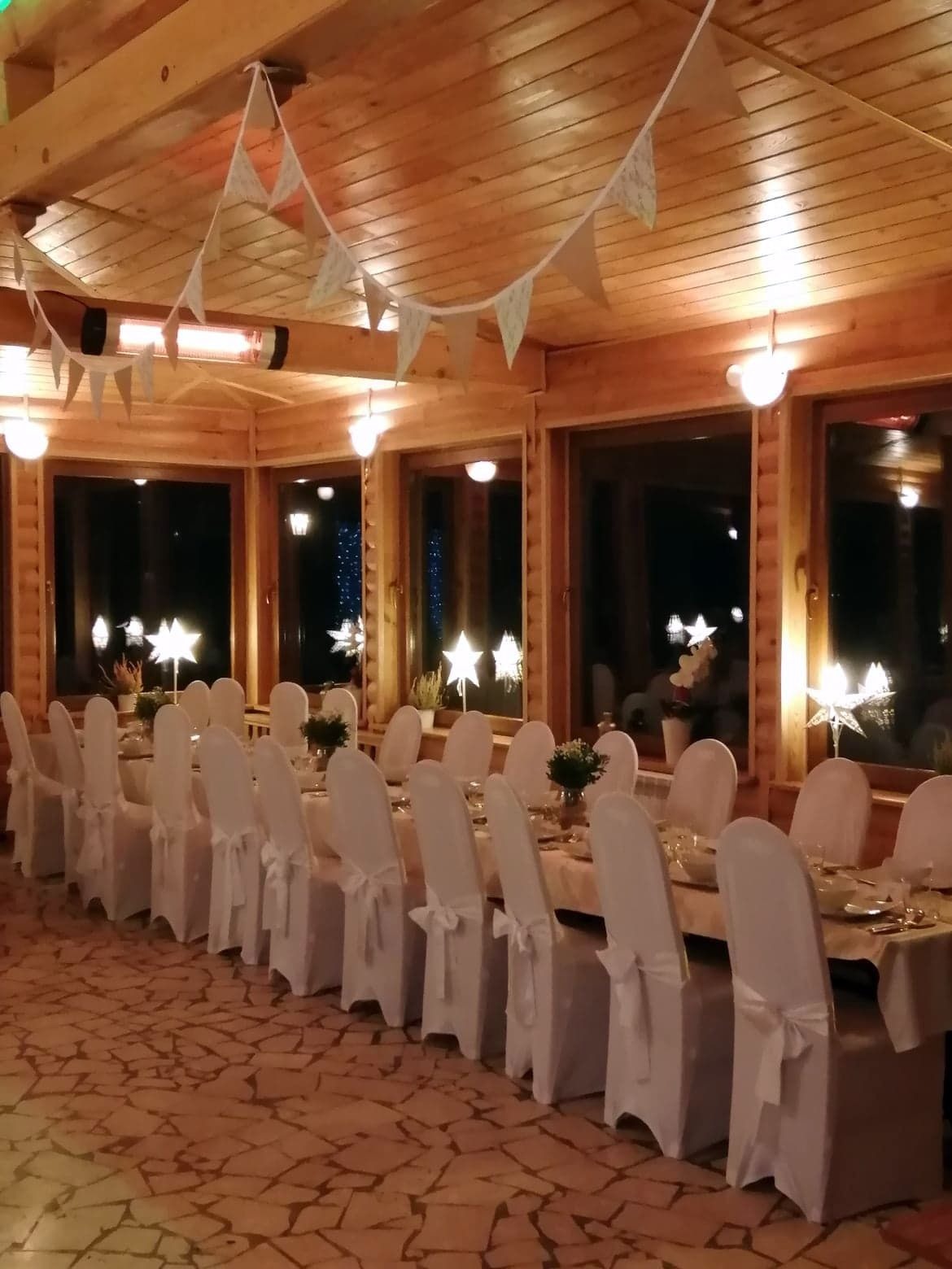 Uroczysta kolacja wigilijna w drewnianej sali, długi stół z białymi krzesłami i dekoracjami gwiazdkowymi, ciepłe oświetlenie i girlanda z chorągiewek.