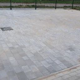 Ułożona szara kostka brukowa na posesji, widoczny fragment buta robotnika i studzienka kanalizacyjna wkomponowana w nawierzchnię.