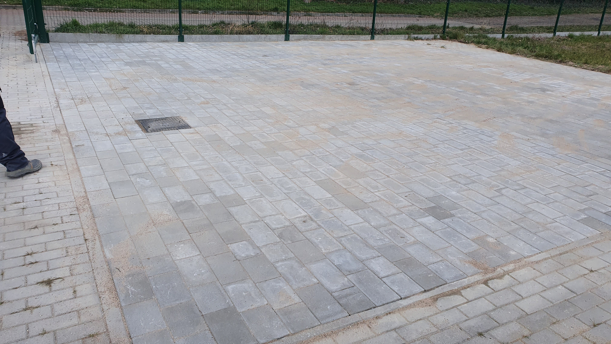 Ułożona szara kostka brukowa na posesji, widoczny fragment buta robotnika i studzienka kanalizacyjna wkomponowana w nawierzchnię.