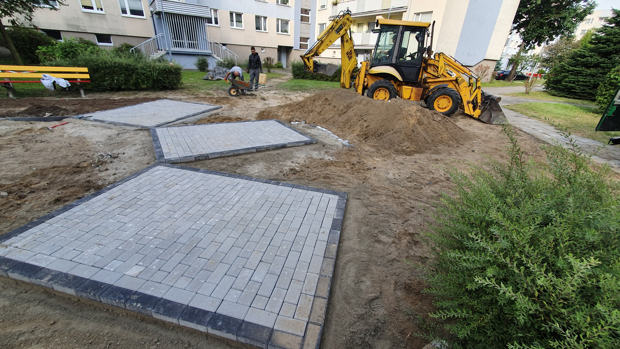 Przygotowanie terenu pod ścieżkę z kostki brukowej: żółta koparko-ładowarka i dwaj pracownicy z taczką na tle bloku mieszkalnego.